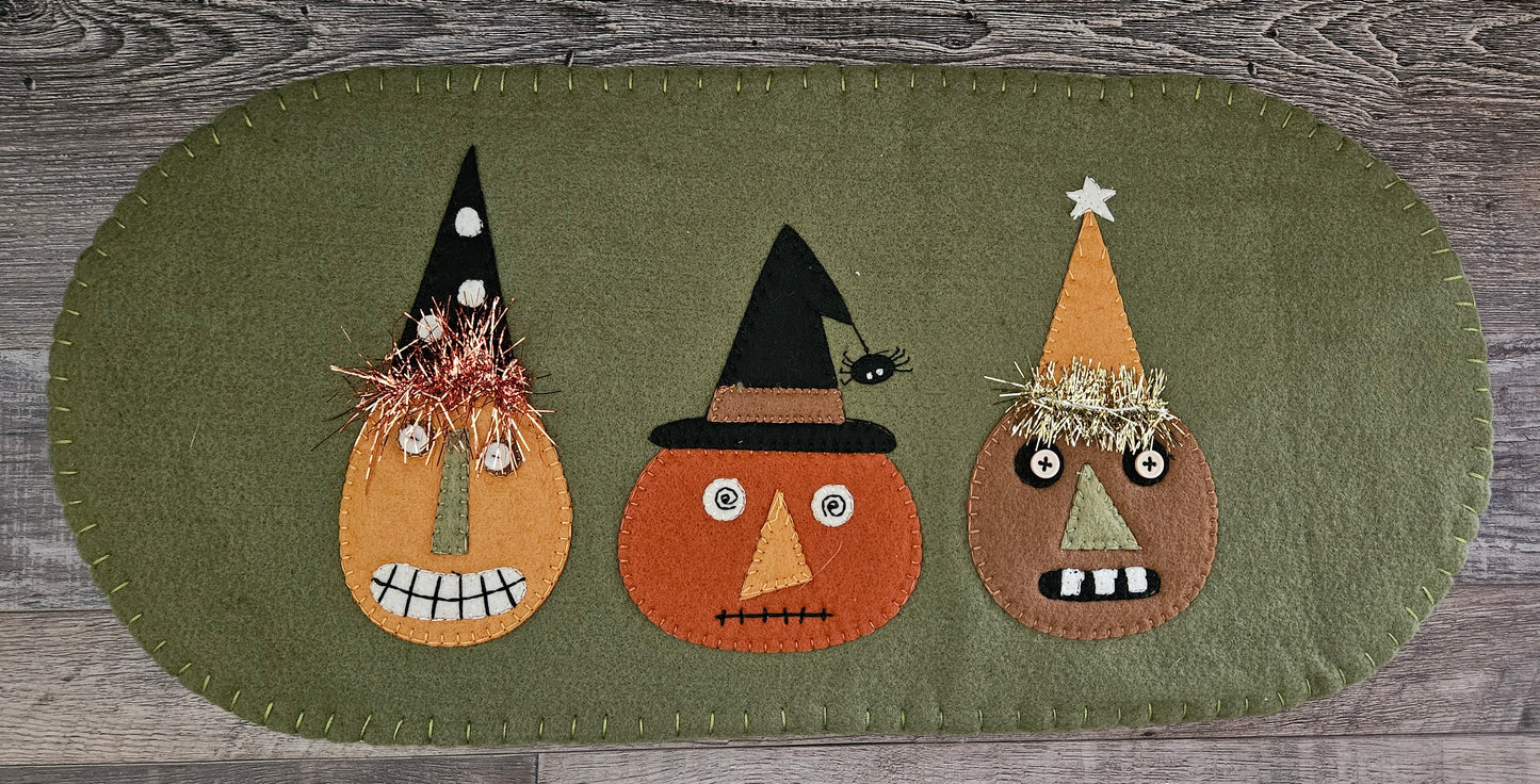 A wool felt table mat featuring three appliqued designs of jack o lanterns with different facial expressions, primarily in brown, orange, and black colors, on a green background.