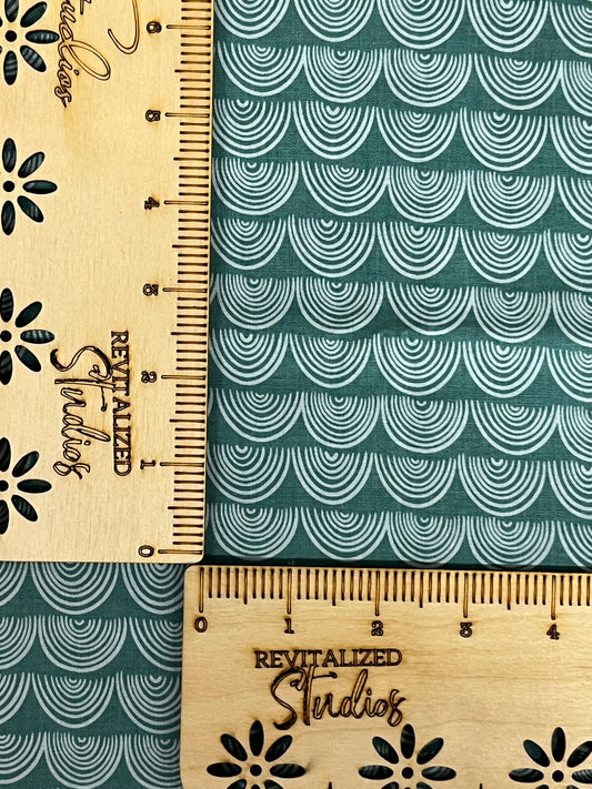 A fabric fat quarter with a teal-green background featuring a wave pattern and the text 'Revitalized Studios' and 'Studied Revitalized'. The ruler on the fabric shows measurements in inches.