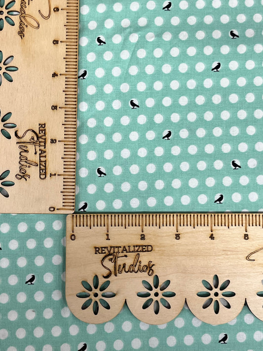 A fat quarter of fabric with a print of black crows on white polka dots against a minty-colored background, with the brand Riley Blake Designs inscribed on the side.