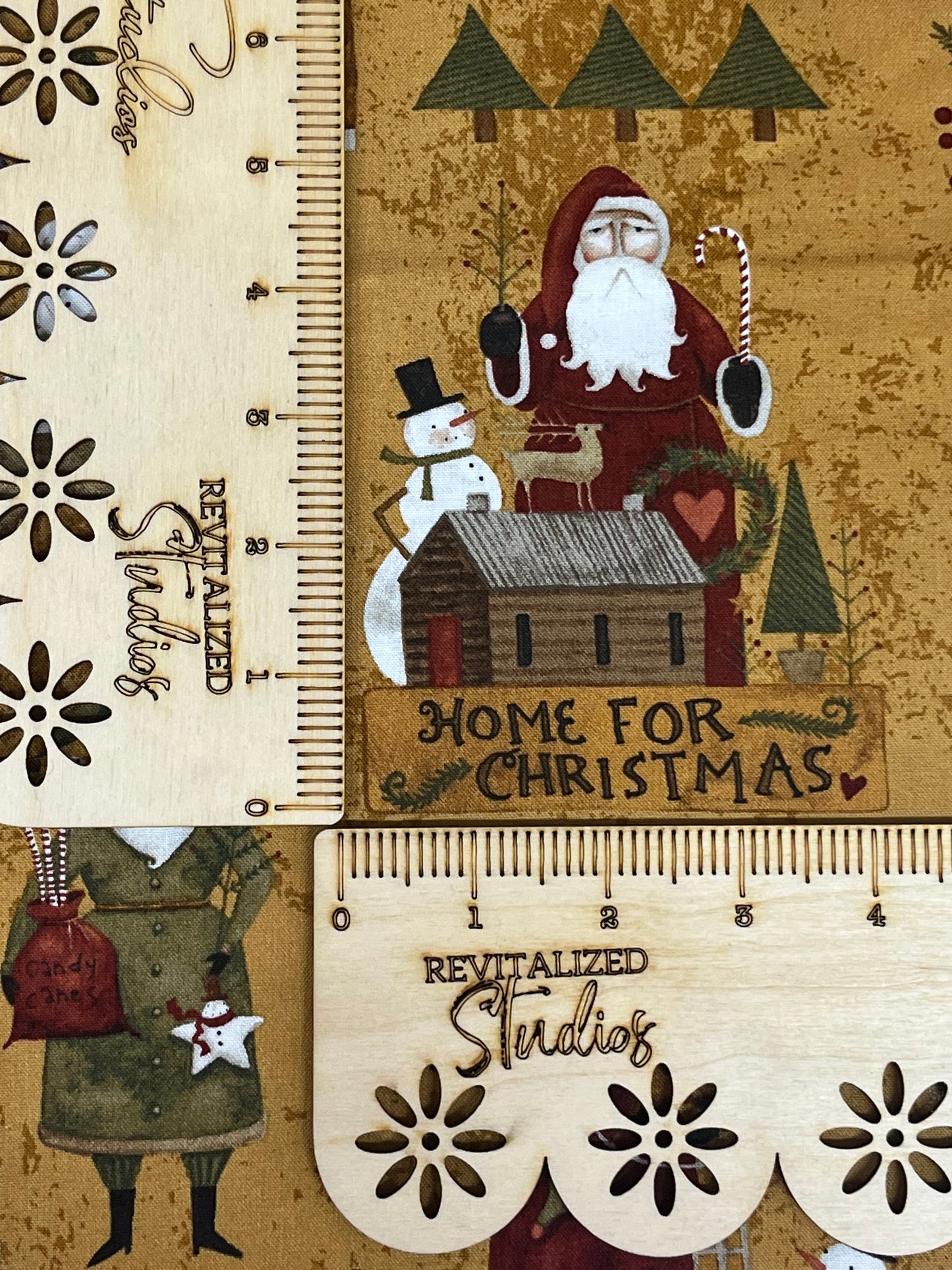 A fabric fat quarter featuring a rustic design with an illustration of Santa Claus, a reindeer, and a Christmas tree on a mustard/brown backdrop. The text on the fabric reads 'Home for Christmas' and 'Revitalized Studios'.